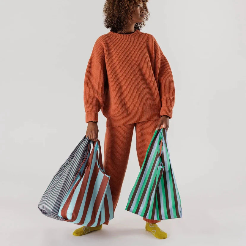 Woman standing holding one brown and blue striped baggu reusable bag and one lilac and blue bag in right hand and one green lilac brown striped baggu reusable bag in left hand