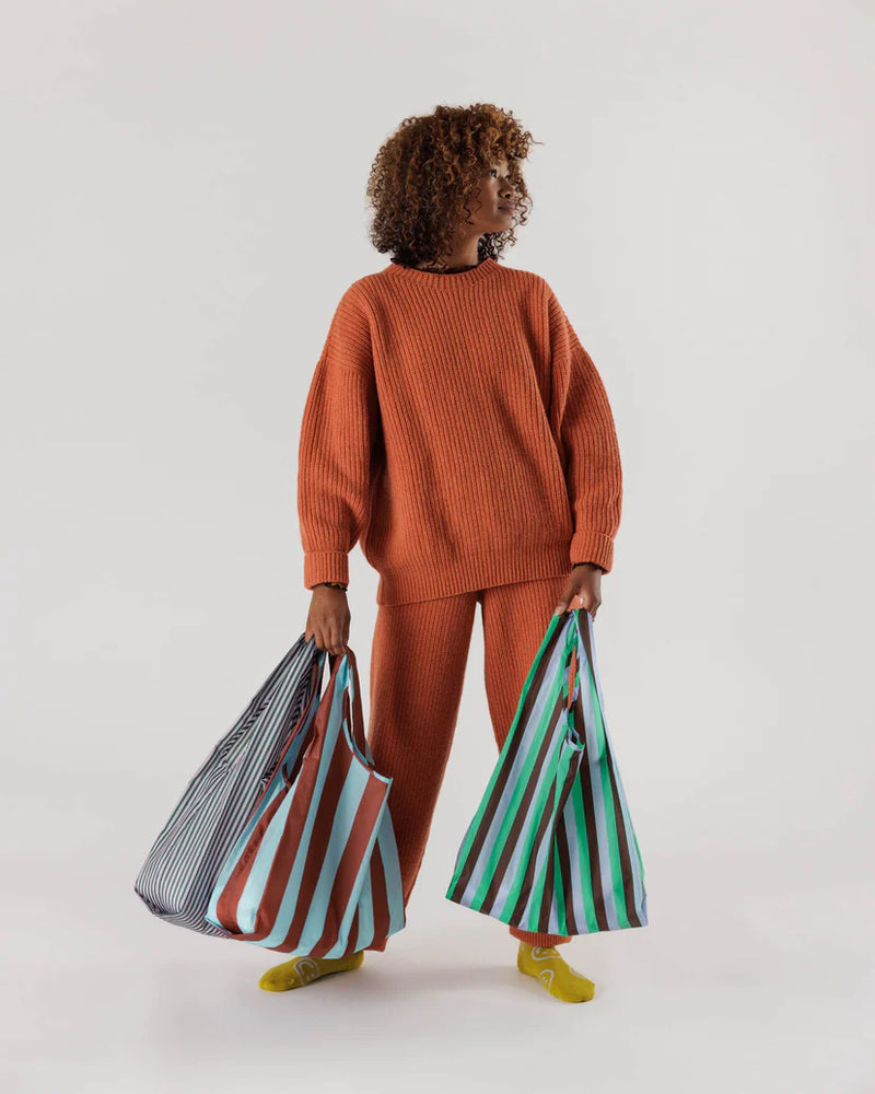 Woman standing holding one brown and blue striped baggu reusable bag and one lilac and blue bag in right hand and one green lilac brown striped baggu reusable bag in left hand
