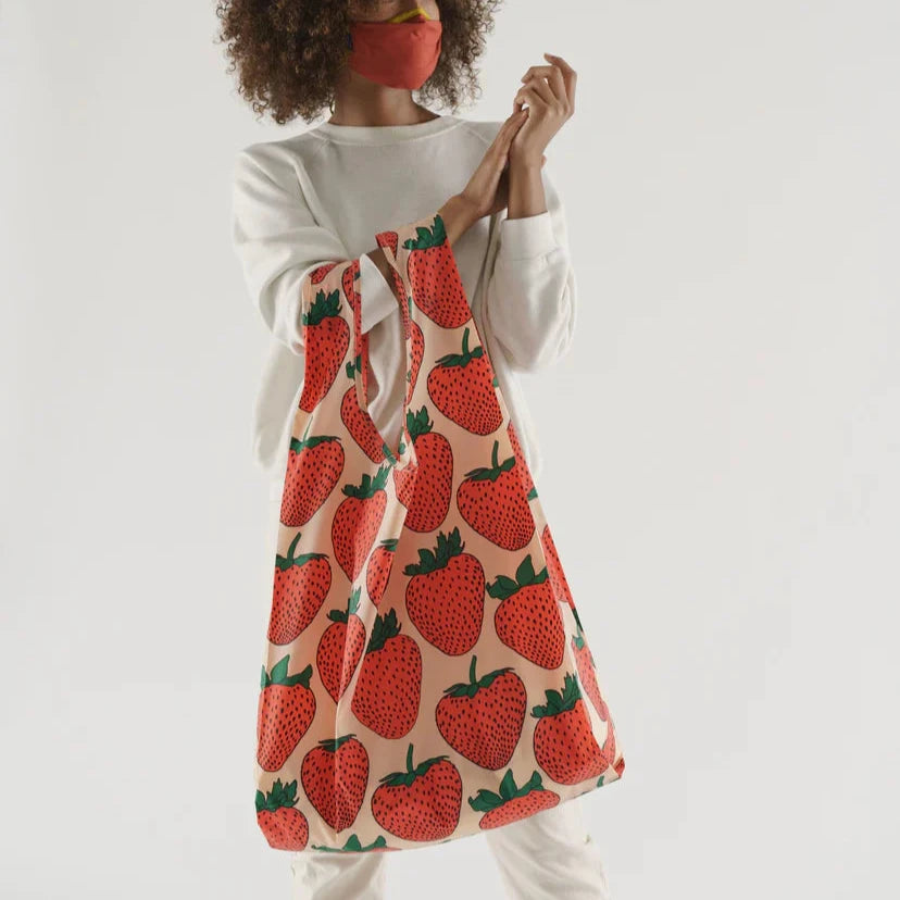 Woman with hands clasped holds both straps on right arm a big baggu designed with red pink and green strawberry pattern open to show full size of reusable bag