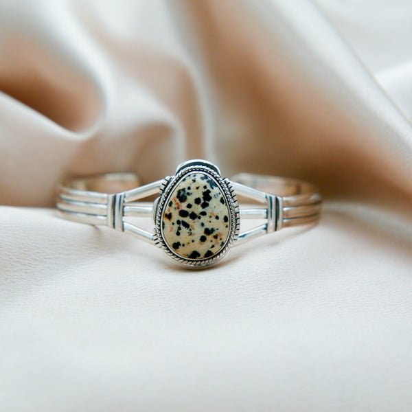 Close up of a sterling silver cuff featuring a teardrop shaped Dalmation Jasper center stone. The stone is bezel set with twisted & stamped silver details. 