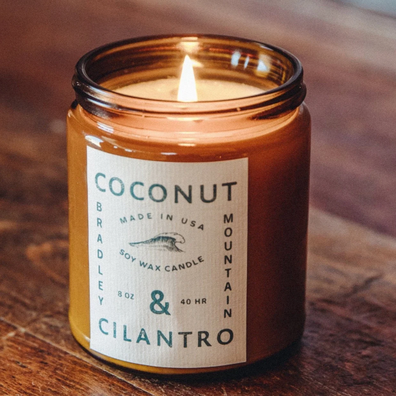 IMage of lit candle on dark would counter, candle labeled "coconut and cilantro bradley mountain" Diameter: 3 in, Height: 3 in Approx. Net Wt: 8 oz Approx. Burn time: 40+ hours Infused with Natural Essential Oils