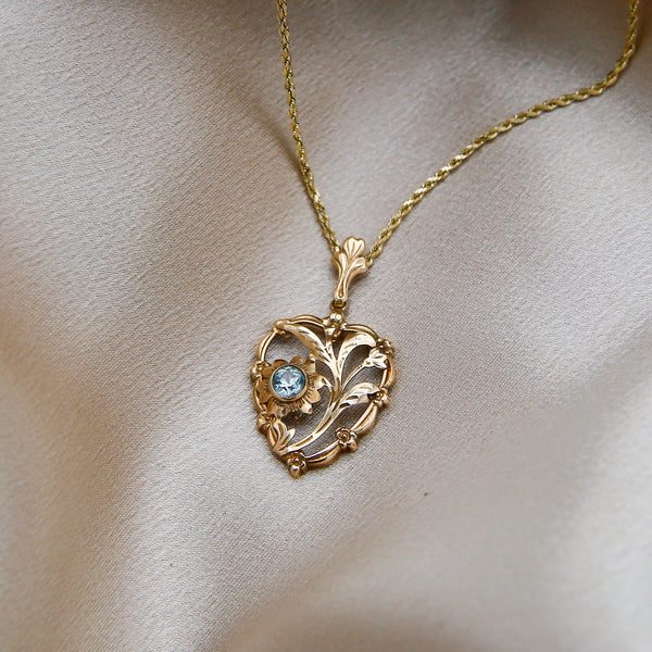 An elaborate heart shaped pendant with floral detail and an aquamarine stone on a gold fill chain. 