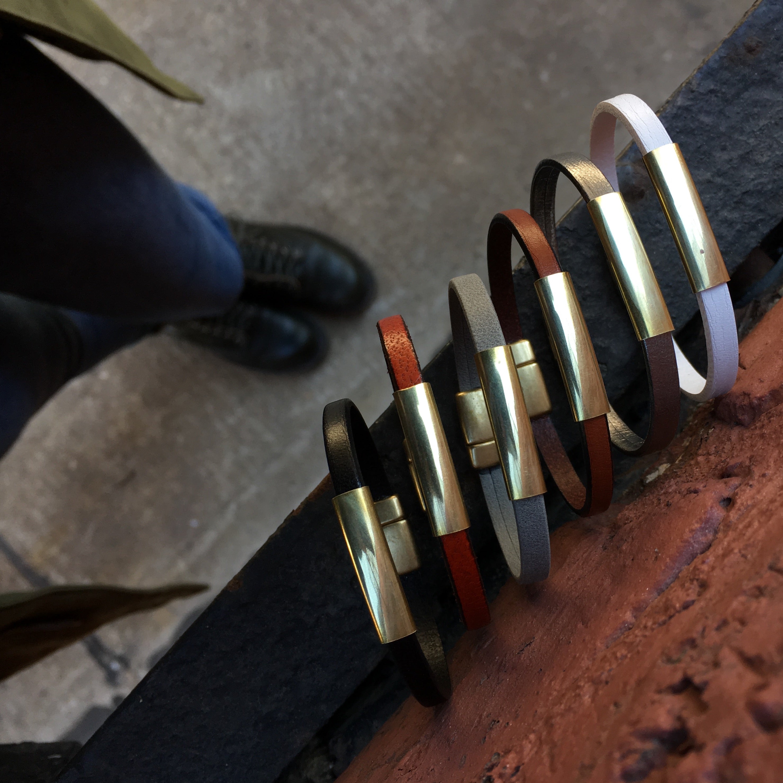 Six "Aida" leather & brass bar bracelets with magnetic clasps in various colors and displayed standing upright on metal beam..