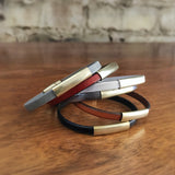 6 "Aida" leather and brass slider bracelets in black, tan, gunmetal, white, and blood orange stacked on a table.