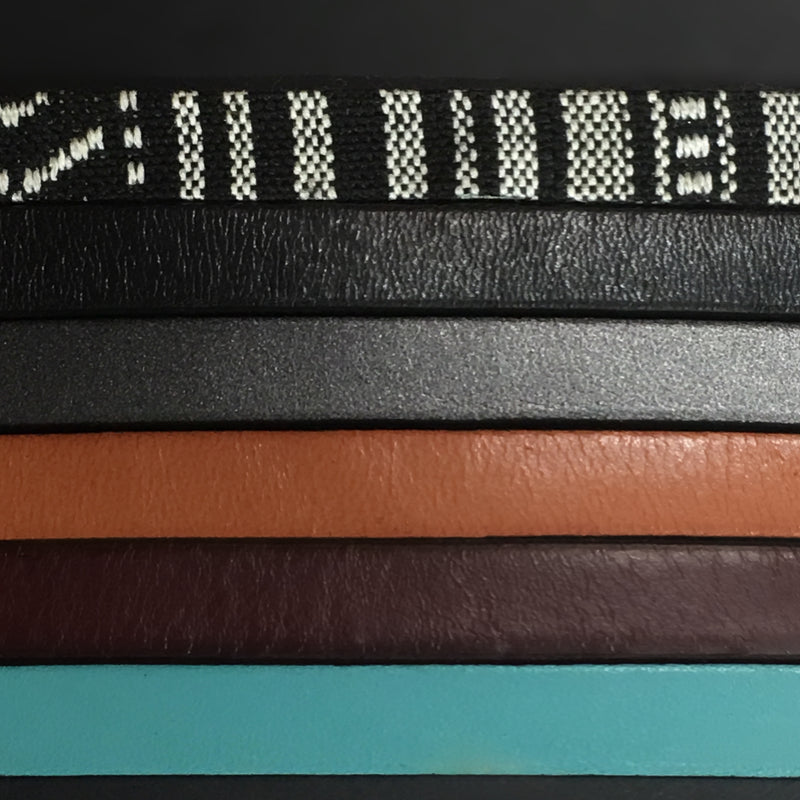 Six strips of leather displaying color options for the "Ace" leather bracelet, the colors are Black & white fabric, Black leather, gunmetal gray leather, tan leather, mahogany leather and turquoise leather.