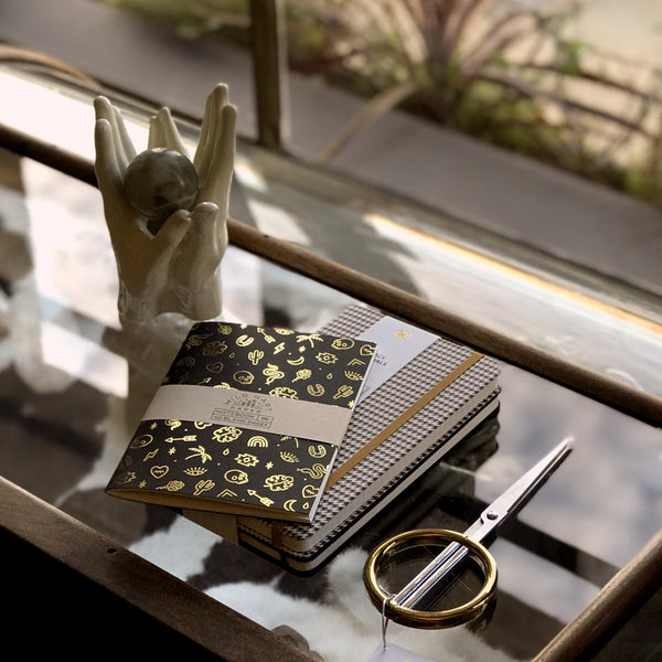 image of notebook on desktop with a pair of scissors and sculptural items. notebook is black with gold foil illustrations. 5"x7"  80# text weight, off white paper Cover is heavy weight black paper stock printed with gold foil. Packaged with labeled belly band in clear cello bag.
