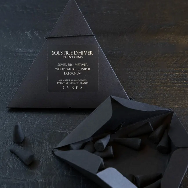 Black paper packaging containing black cone incense spilled out onto dark table. Product labeled "solstice d'hiver' incense cones. Scent siver fir, vetiver, wood smoke, juniper, labdanum
