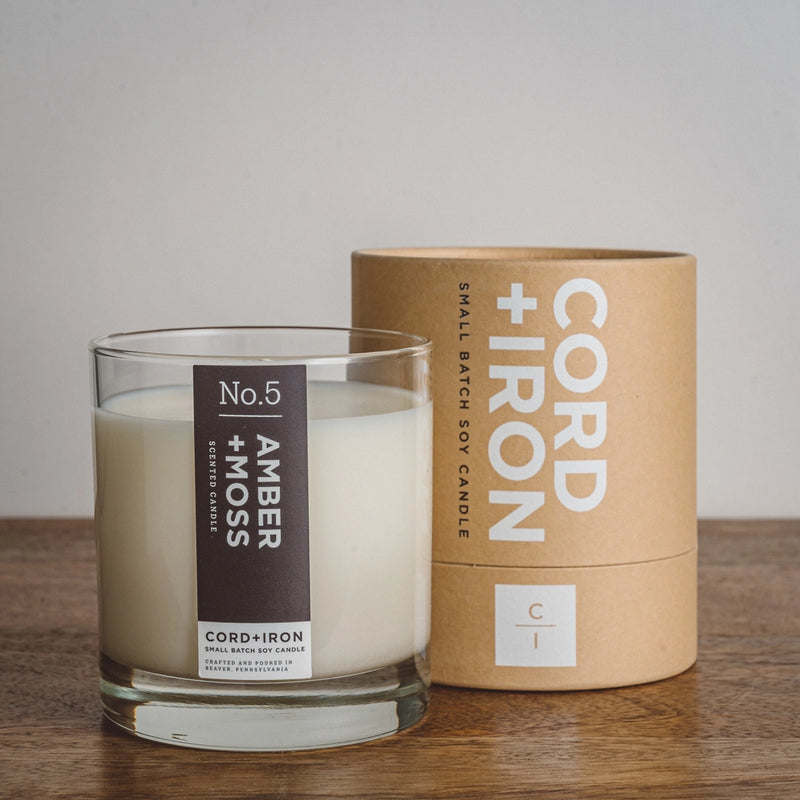 Image of candle and tube container on dark wood counter, labeled "amber and moss no. 5" by cord + Iron small batch soy candle 