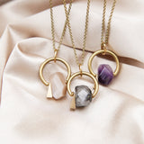 Three variants of a suspension necklace with brass components. One with a peach moonstone, one with tourmalinated quartz and one with an amethyst gemstone.  