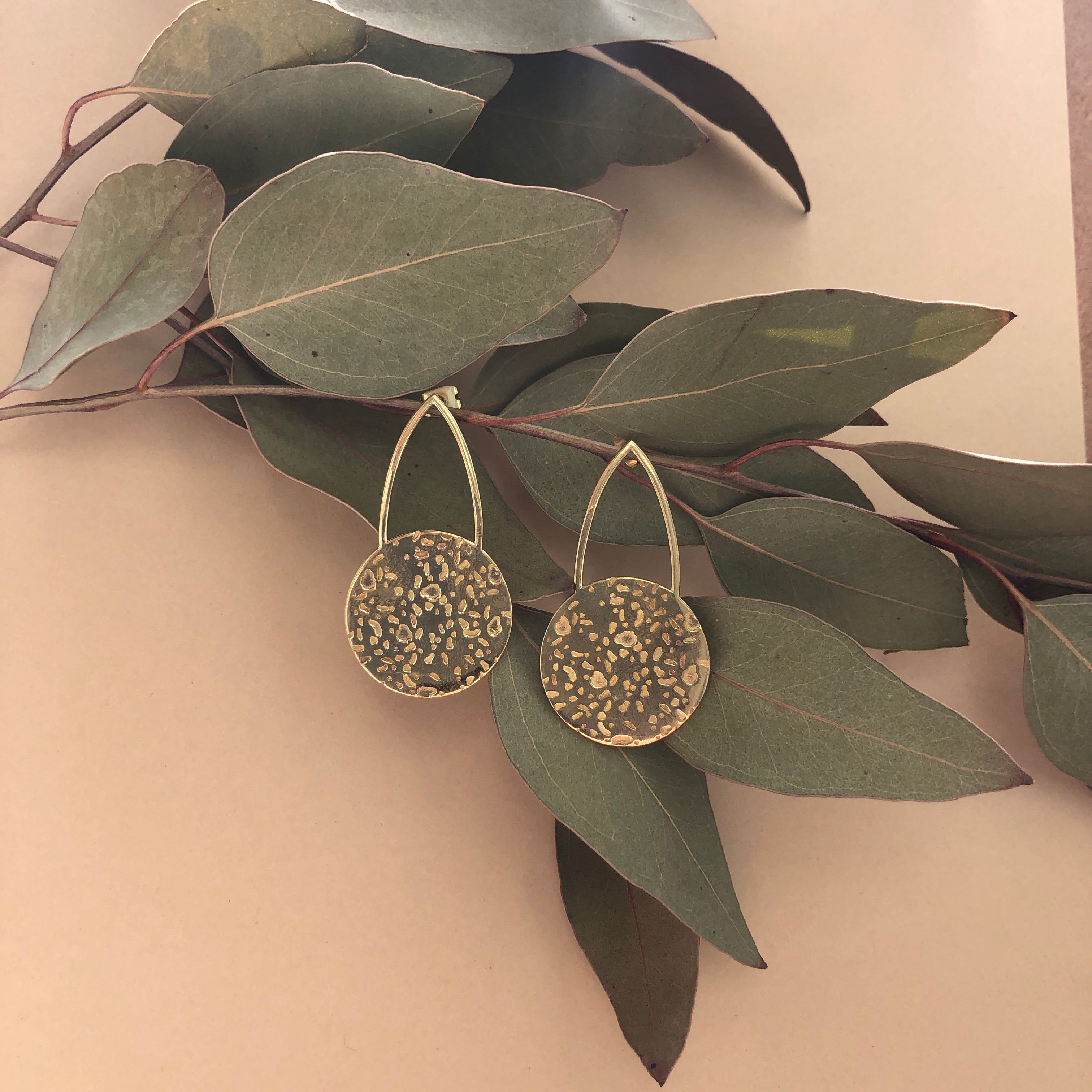 Pair of "Gwen" large brass cast teardrop stud earrings that have a textured brass circle at the bottom of the open teardrop shape.