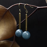 Close up of "Marit" drop earrings that have a vertical brass bar with a blue angelite round below.