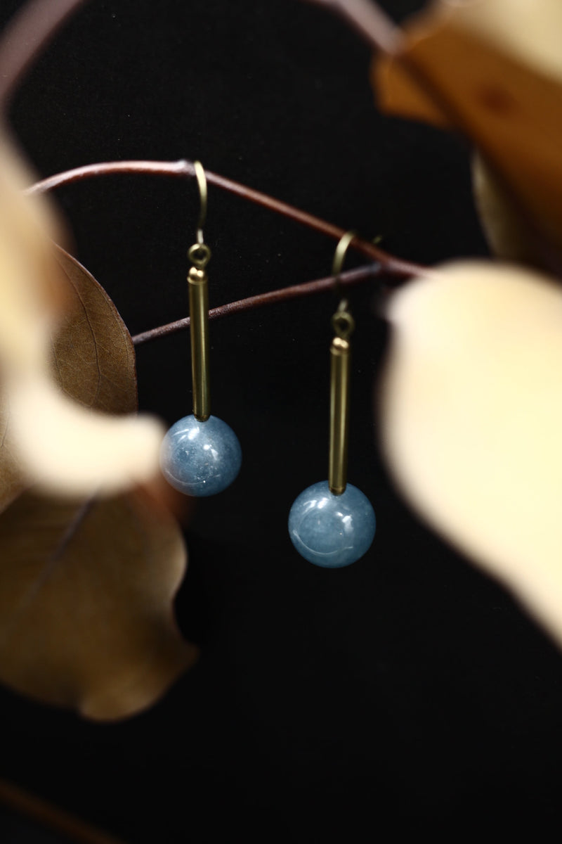 Close up of "Marit" drop earrings that have a vertical brass bar with a blue angelite round below and are hanging on ear wires.
